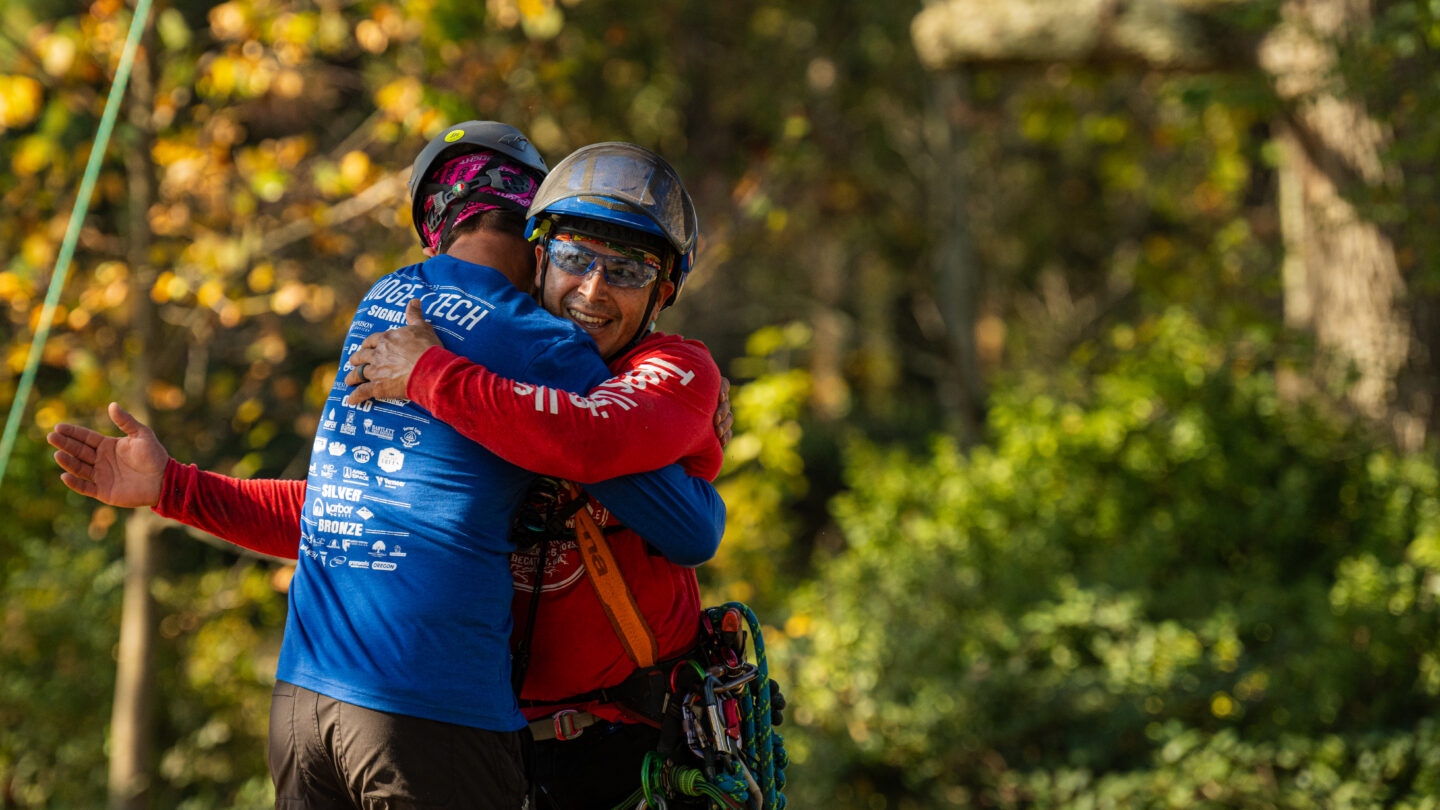 A Man in a blue shirt embraces a man in a red shirt who is wearing a helmet, safety goggles, and a gear belt clipped into climbing ropes.