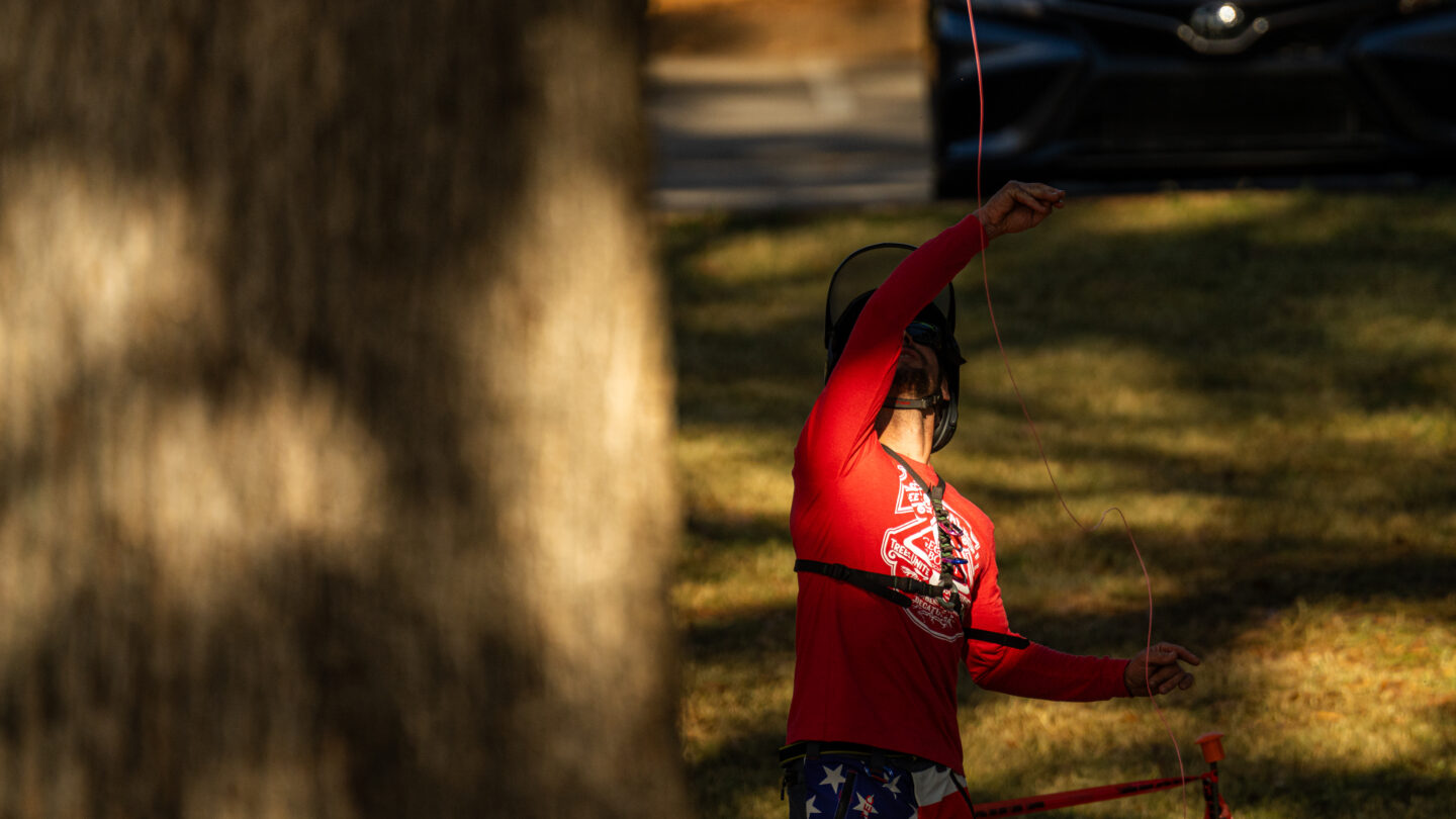 A man in a red shirt and American-flag print pants hoists a red rope over a tree limb.