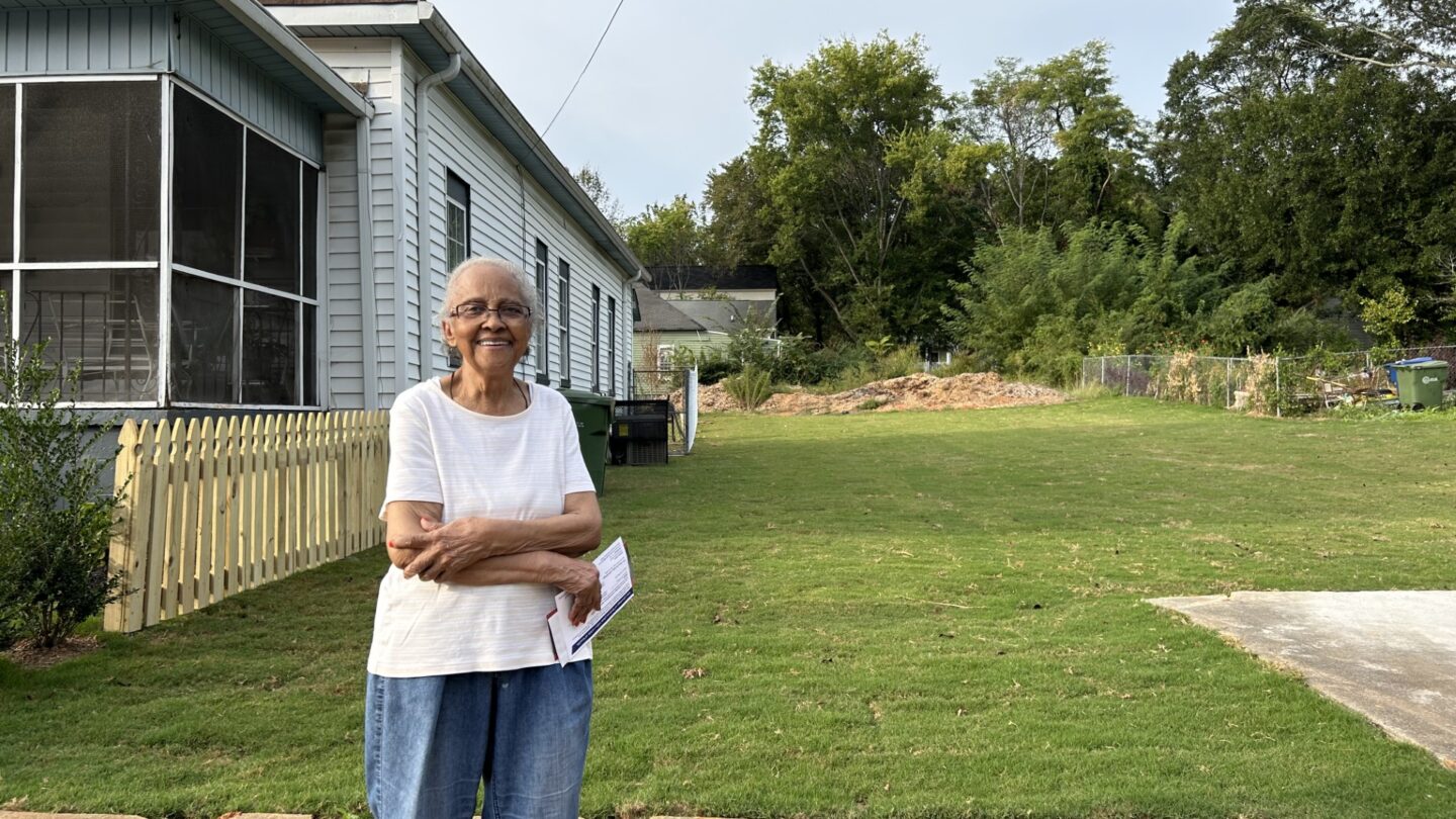 An older lady stands in front of a short, stone wall and green lawn laid with fresh sod. Her house is to the left, a gray one-story with a short wood picket fence and screened-in porch.