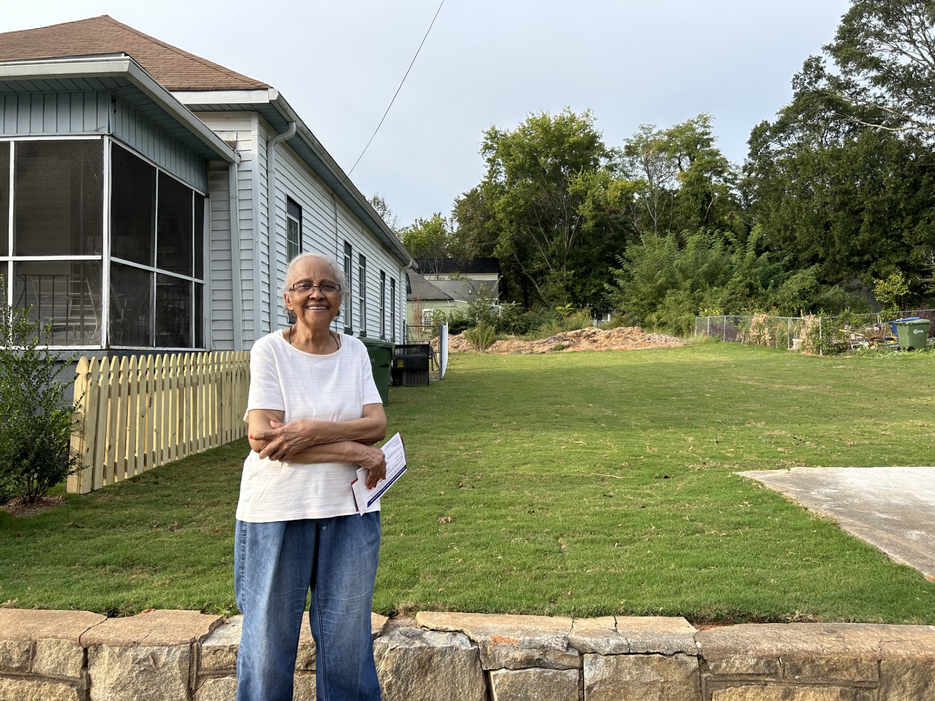 An older lady stands in front of a short, stone wall and green lawn laid with fresh sod. Her house is to the left, a gray one-story with a short wood picket fence and screened-in porch.