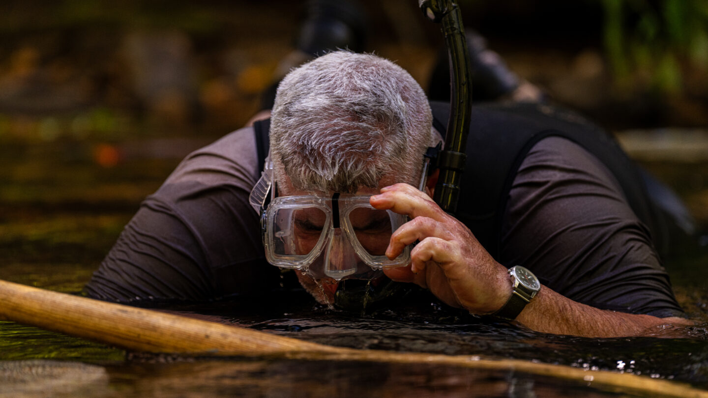 A man wearing goggles lays on his chest flat in a shallow stream.