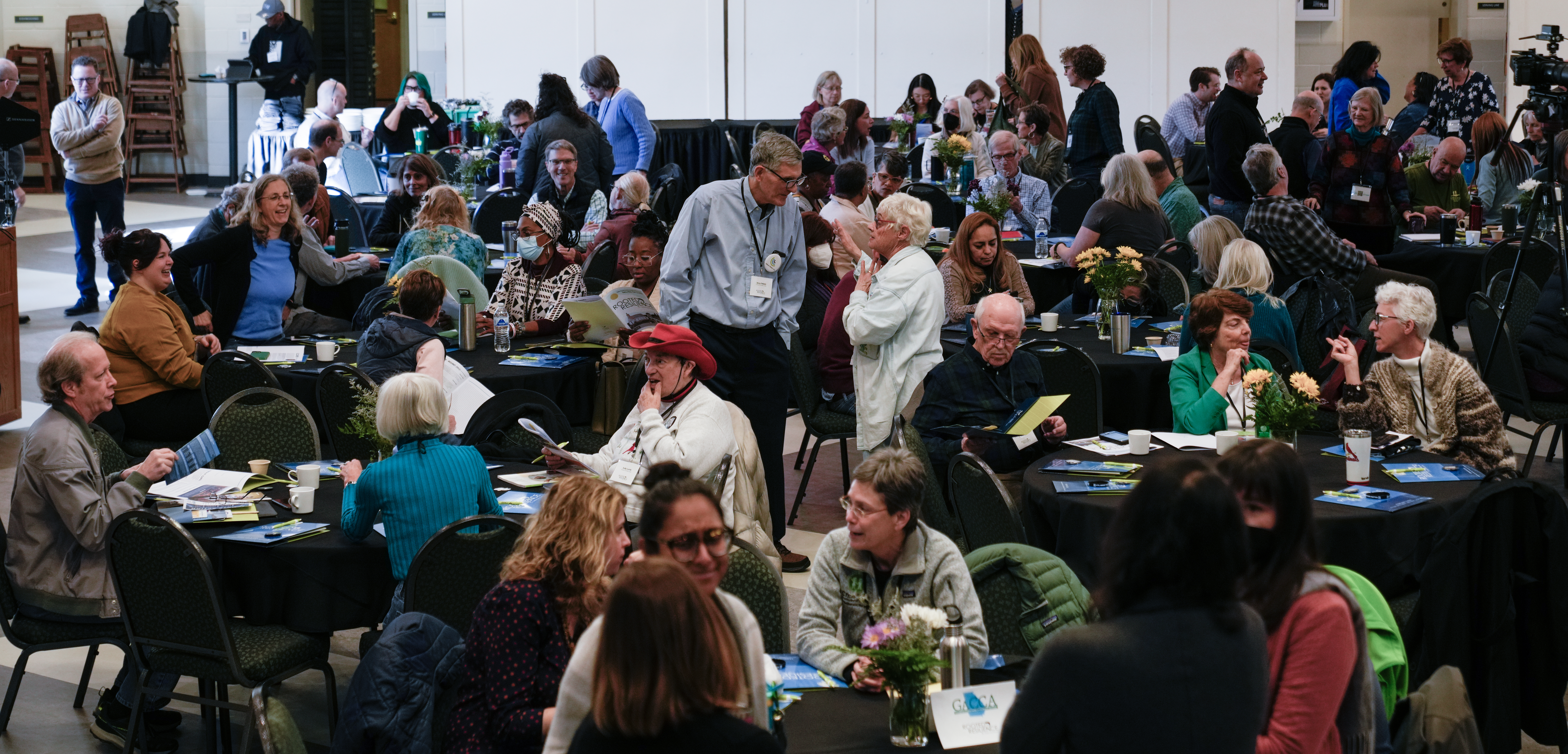 A crowd of people sit and stand around circular tables at a summit for faith-based environmental groups.