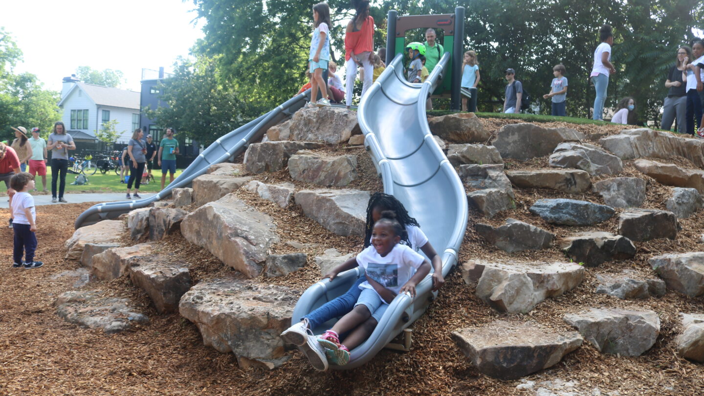 Children slide down a gray slide surrounded by rocks at a park.