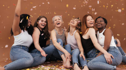 Group of happy women sitting on the brown background while petals from flowers falling. Laughing females of different races and ages under petals from flowers.