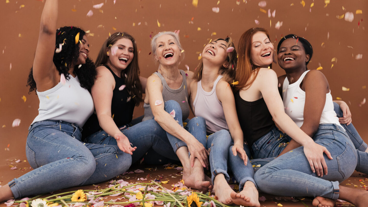 Group of happy women sitting on the brown background while petals from flowers falling. Laughing females of different races and ages under petals from flowers.