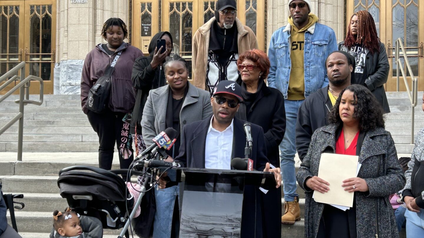 Mahdi Chaney, wearing a Braves baseball cap, sunglasses and sports coast speaks at a podium on the steps of City Hall next to his wife. She is wearing a gray peacoat with her hand on a stroller containing her two children.