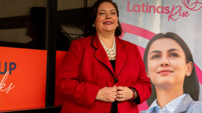 A woman wearing a red blazer standing in front of a banner that reads Latinas Rise.
