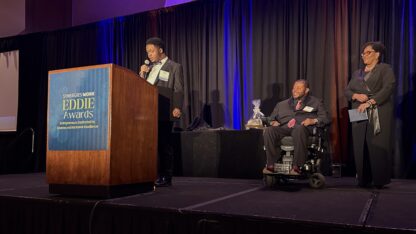 Three people, with one person in a wheelchair are on stage at an awards gala.