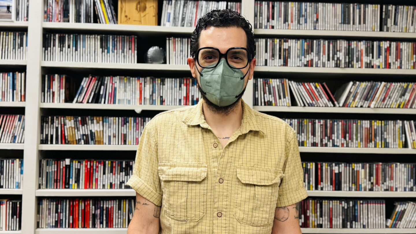 Dom Kelly, co-founder of New Disabled South, wears a mask and glasses and stands in front of a bookshelf at the WABE studio.