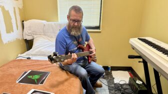 Charles Walsh sits on his bed and plays the bass guitar inside his room at CaringWorks Hope House.