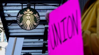 Cassandra Gonzalez holds a union sign while passing in front a Starbucks logo as workers participate in a walkout and strike during the company's Red Cup Day Thursday, Nov. 16, 2023, near Pike Place Market in Seattle.