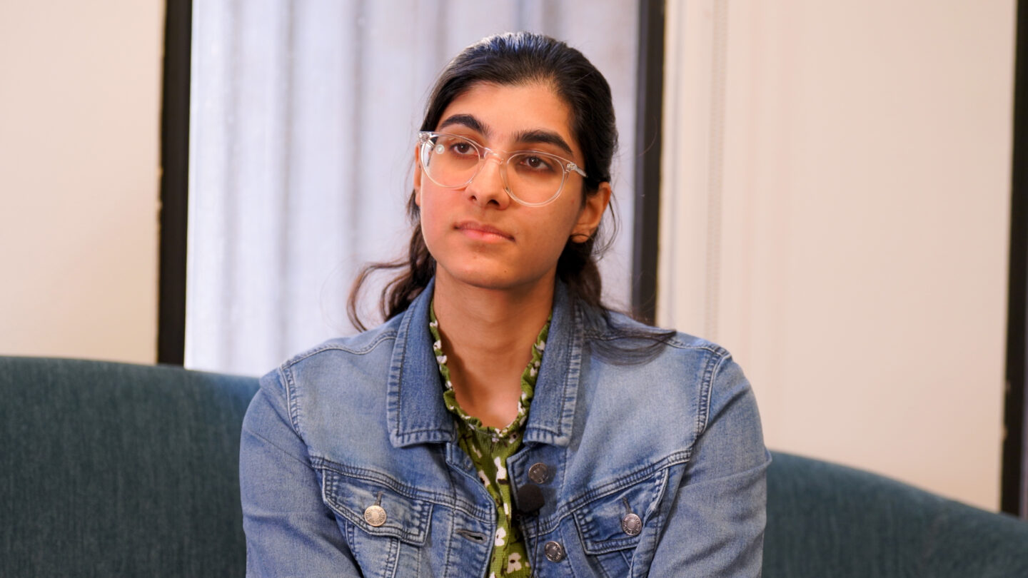 A woman wearing glasses and a jean jacket sits on a couch, looking off to the left.