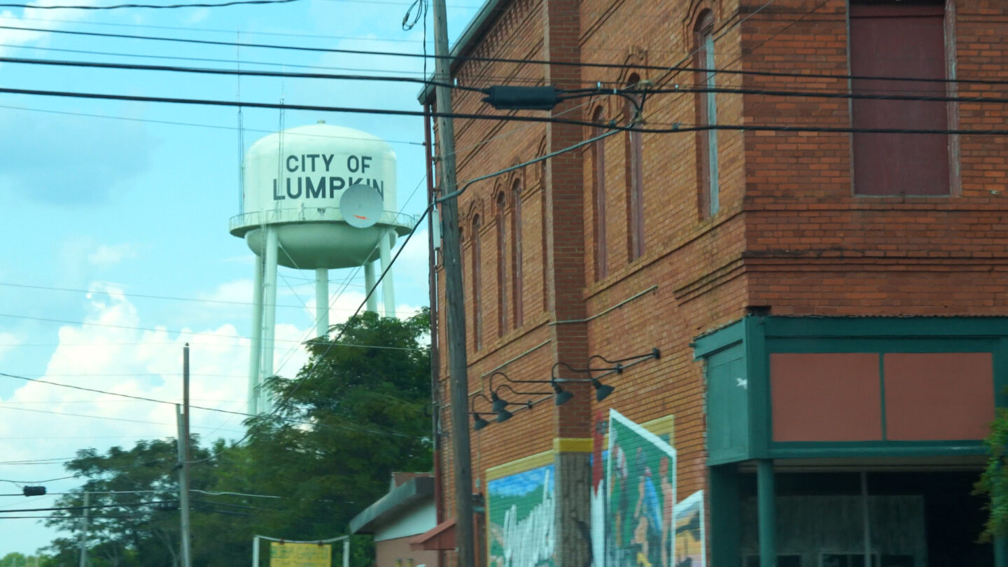 A water tower with black lettering that says City of Lumpkin, next to a brick building in Lumpkin, Georgia.