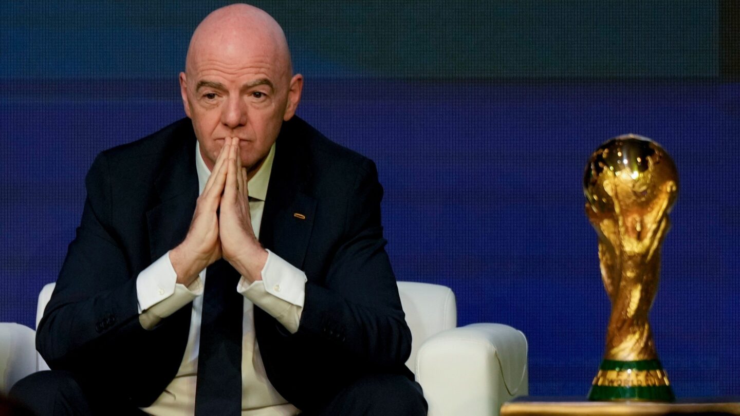 A man in a suit sits in a white chair next to the World Cup trophy