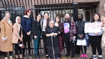 A group of people stand in front of a black wire gate outside a detention facility.