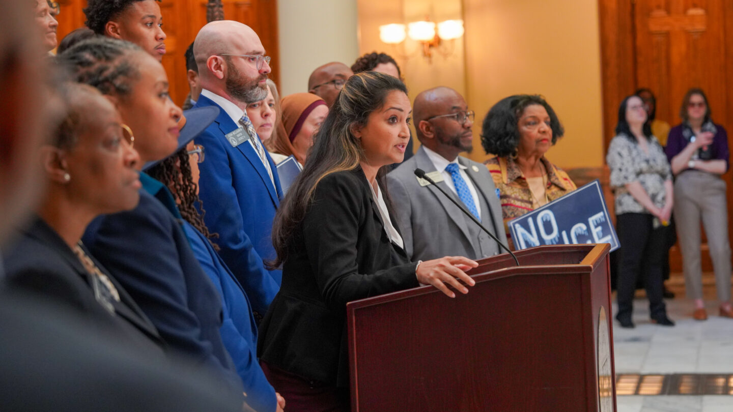 A senator stands at a podium and speaks into a microphone while being surrounded by other lawmakers