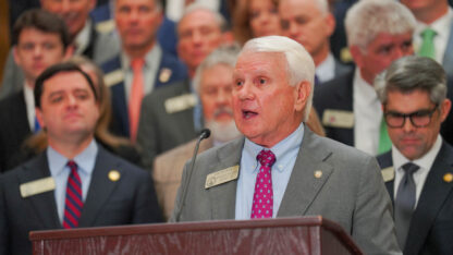 Jon Burns speaks at a podium at a news conference. Behind him is a crowd of other lawmakers.