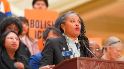 A lawmaker speaks at a podium while others stand behind, holding signs that say Abolish ICE