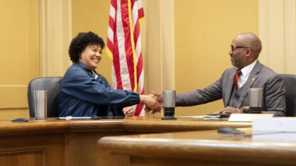 A woman and a man shake hands behind a desk in front of the American flag