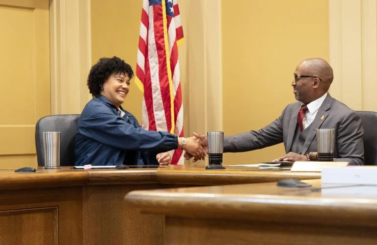 A woman and a man shake hands behind a desk in front of the American flag