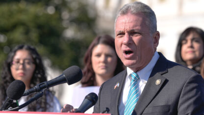 A man in a blue tie and suit speaks at a microphone with three people behind him
