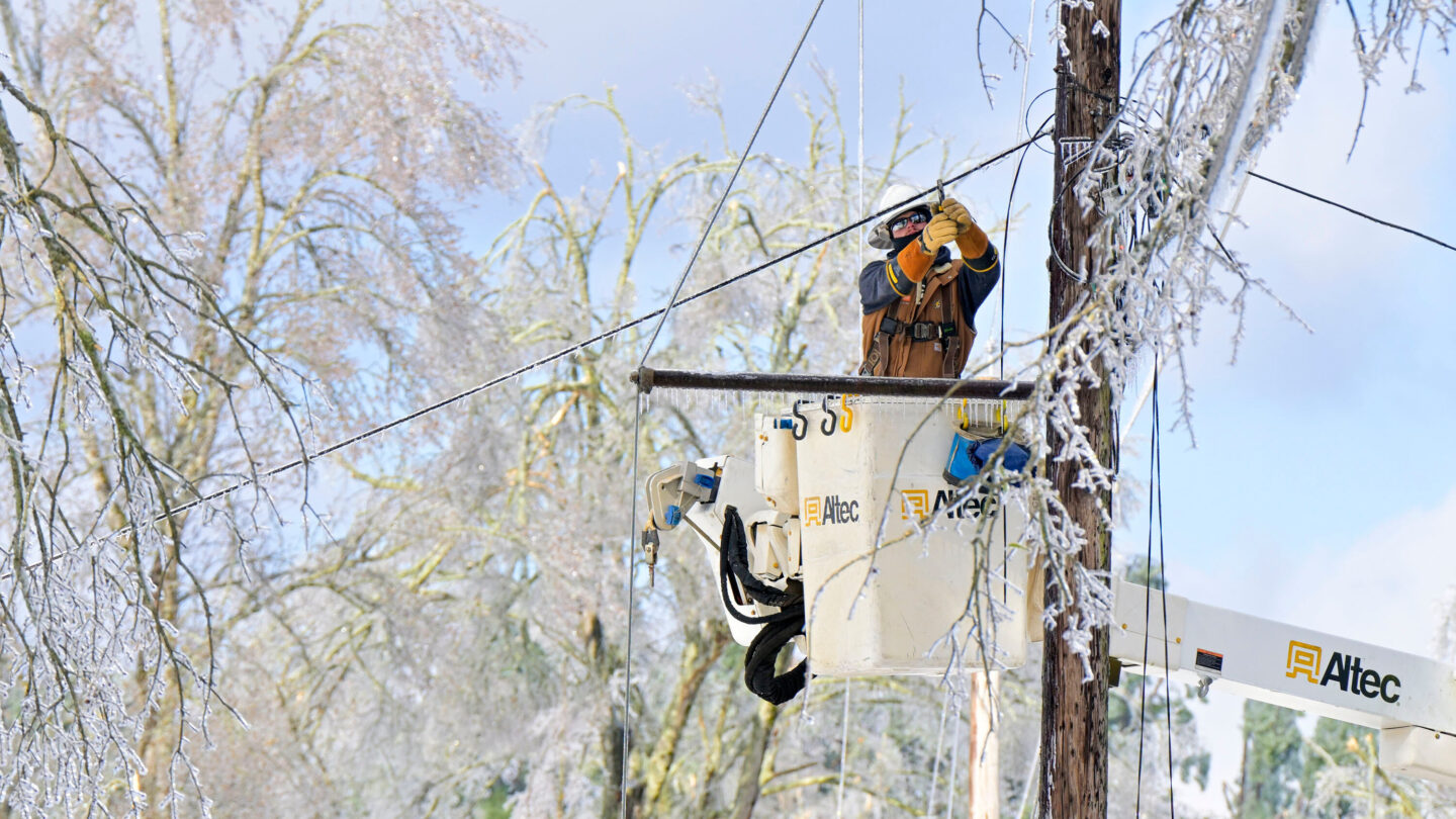 A power lineman works on fixing a power line as ice covers trees behind him