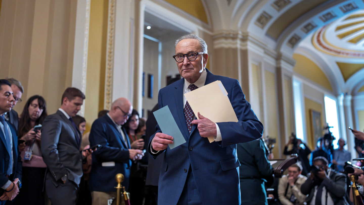 A man in glasses holding a folder walks down a hallway filled with people