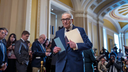 A man in glasses holding a folder walks down a hallway filled with people