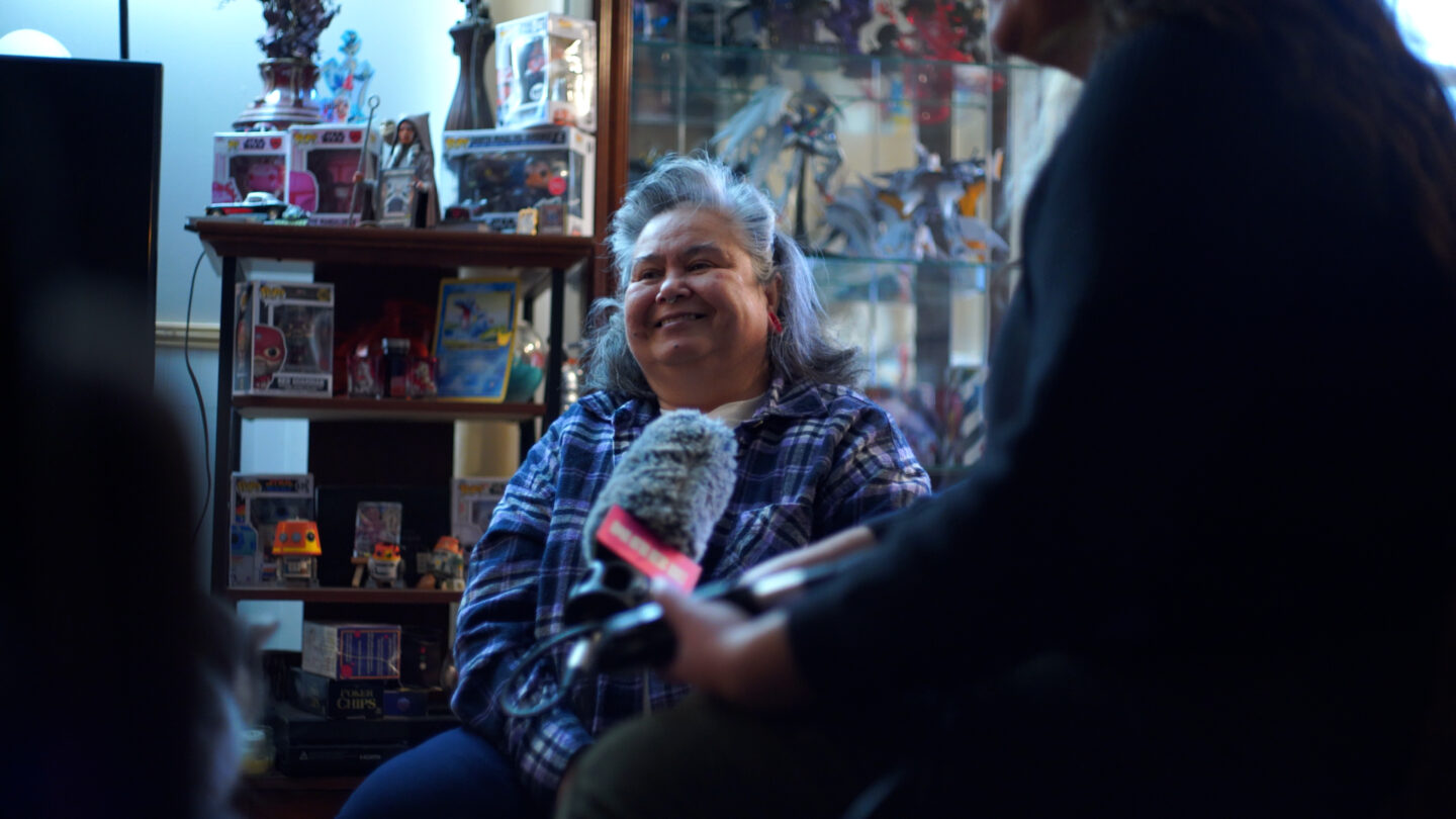 A woman sits in front of several shelves, smiling. A reporter holding a microphone is in the foreground.