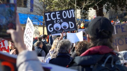 A person holds a large black posterboard with the words ICE OUT FOR GOOD, the two Os in GOOD replaced with giant googly eyes.