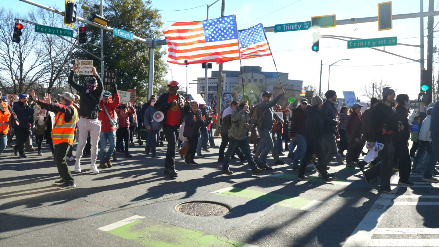 A group of people holding signs and flags march down a street in a protest