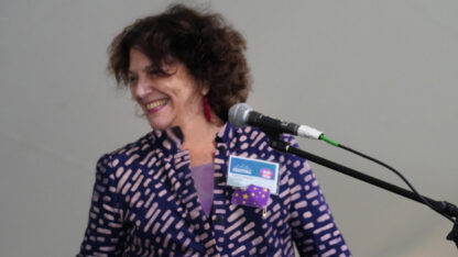 A women in purple jacket with a name tag stands behind a microphone and smiles.