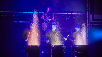 Rockstar character performs electronic percussion beneath illuminated tubes