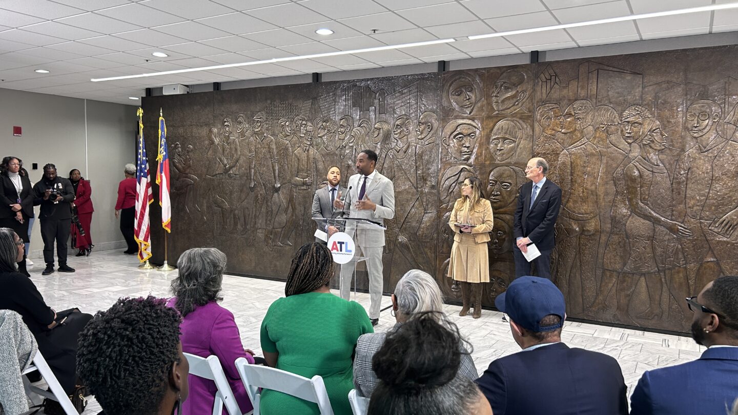 Four people, including the mayor of Atlanta, stand in front of a crowd of people inside a room at Atlanta City Hall