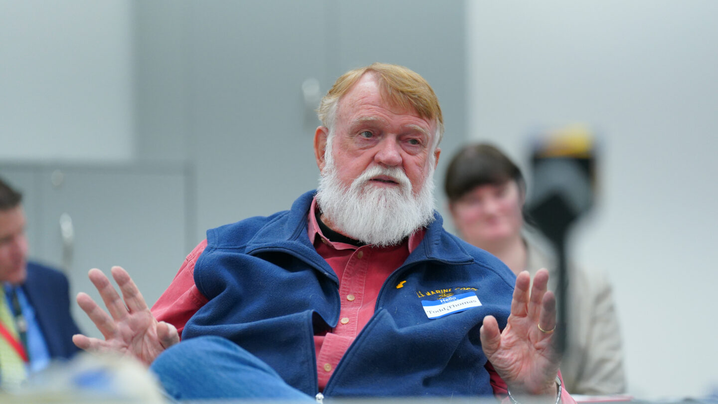 A man wearing a vest and a salmon button down shirt speaks while sitting at a white table