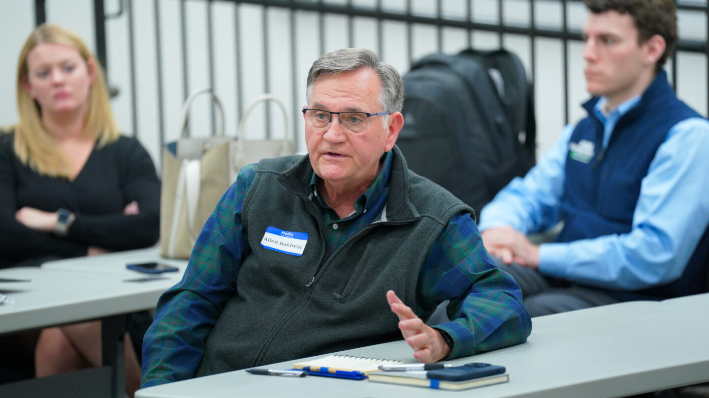 A man sits at a table and speaks whle two other people sit near him