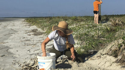 A person kneels on the beach by a bucket in the foreground, while a person stands behind them in the background