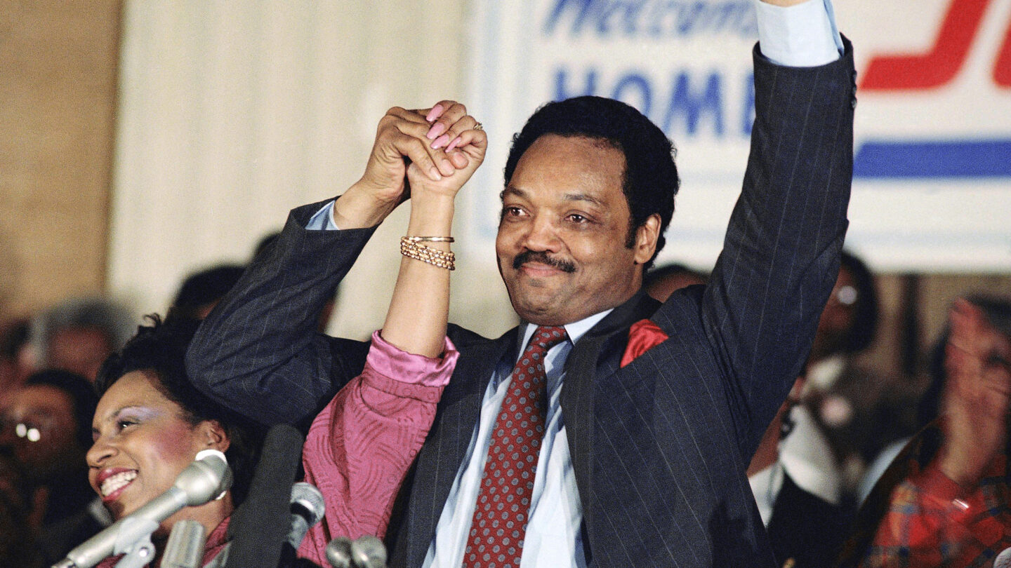 Jesse jackson holds his wife's hand in celebration in front of a cheering crowd