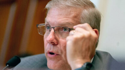 Barry Loudermilk holds his glasses while speaking into a microphone