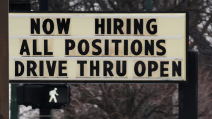 A sign with black letters on a white background says that a business is hiring