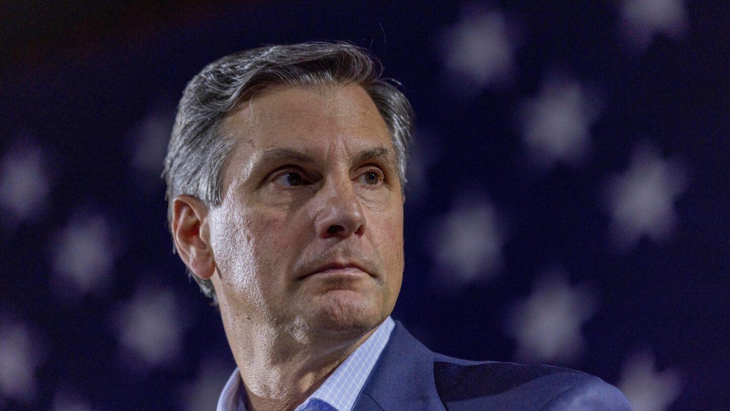 A man in a business suit stands in front of an american flag and looks off to his left