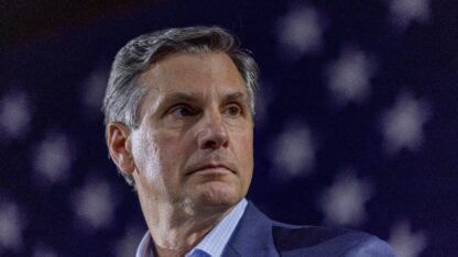 A man in a business suit stands in front of an american flag and looks off to his left