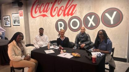 A group sits at a table in front of a Coca Cola ROXY mural