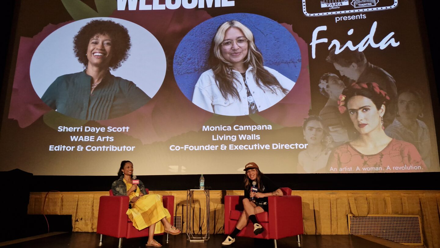 Sherri Daye Scott and Monica Campana sit on stage at the Plaza Theatre in Atlanta beneath a welcome slide showing their names.