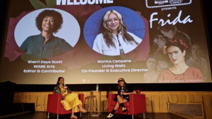 Sherri Daye Scott and Monica Campana sit on stage at the Plaza Theatre in Atlanta beneath a welcome slide showing their names.