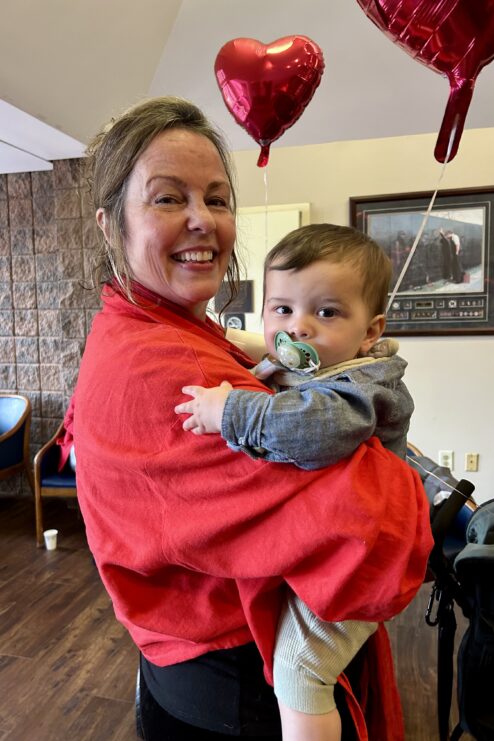 woman in a red jacket holds a baby with heart balloons in the background.