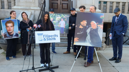 A woman speaks at a press conference surrounded by large posterboard pictures and other people.