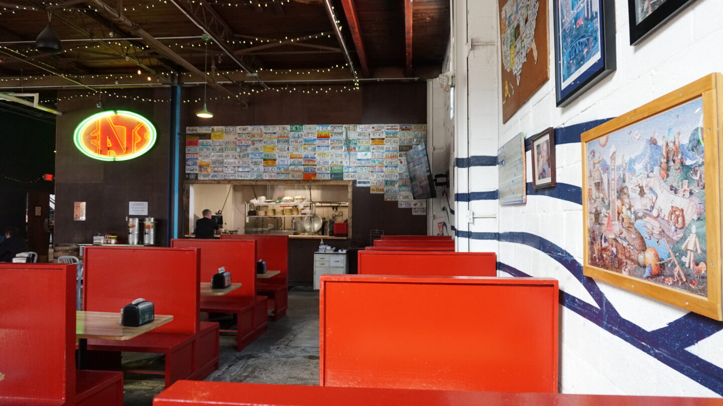 Red wooden booths in front of a wall lined with license plates and a neon EATS sign.