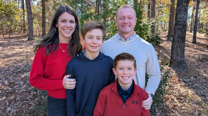 A family four, with a mother, father, and two sons poses for a photo in the woods