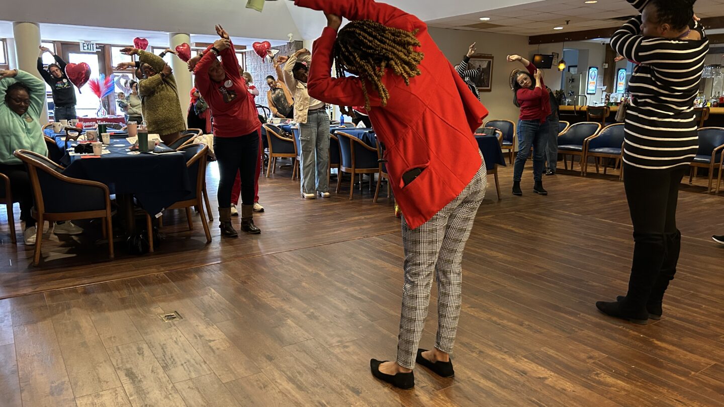 Women stand in a line with their arms over their heads doing warm-up stretches.